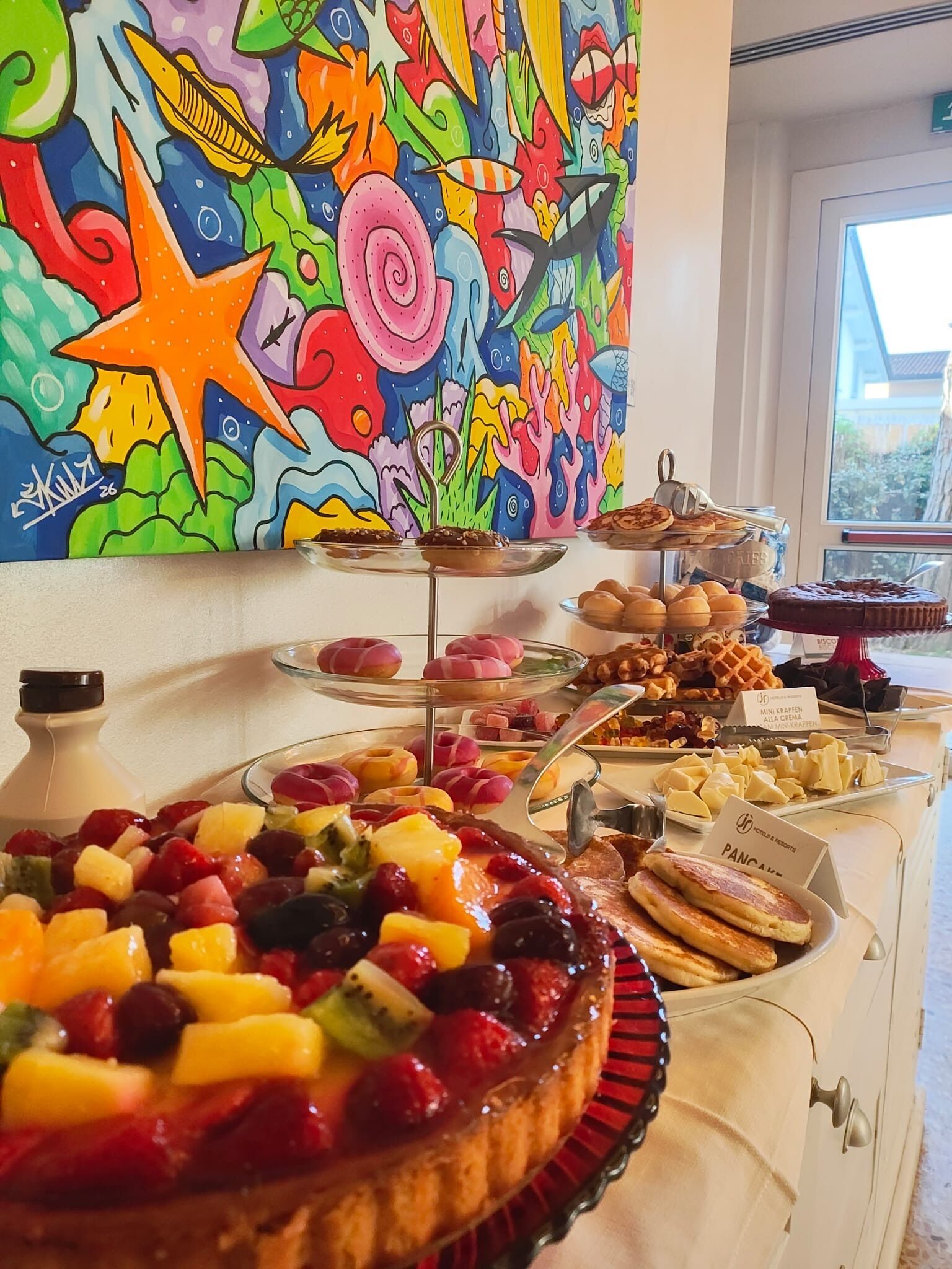 A buffet with a variety of desserts and fruits, a colorful mural on the wall, and a window with a view of the outside.