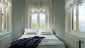 A bedroom with a bed, two bedside tables, a window with a view of the sea, and white curtains. - Camin Hotel Luino (Luino)