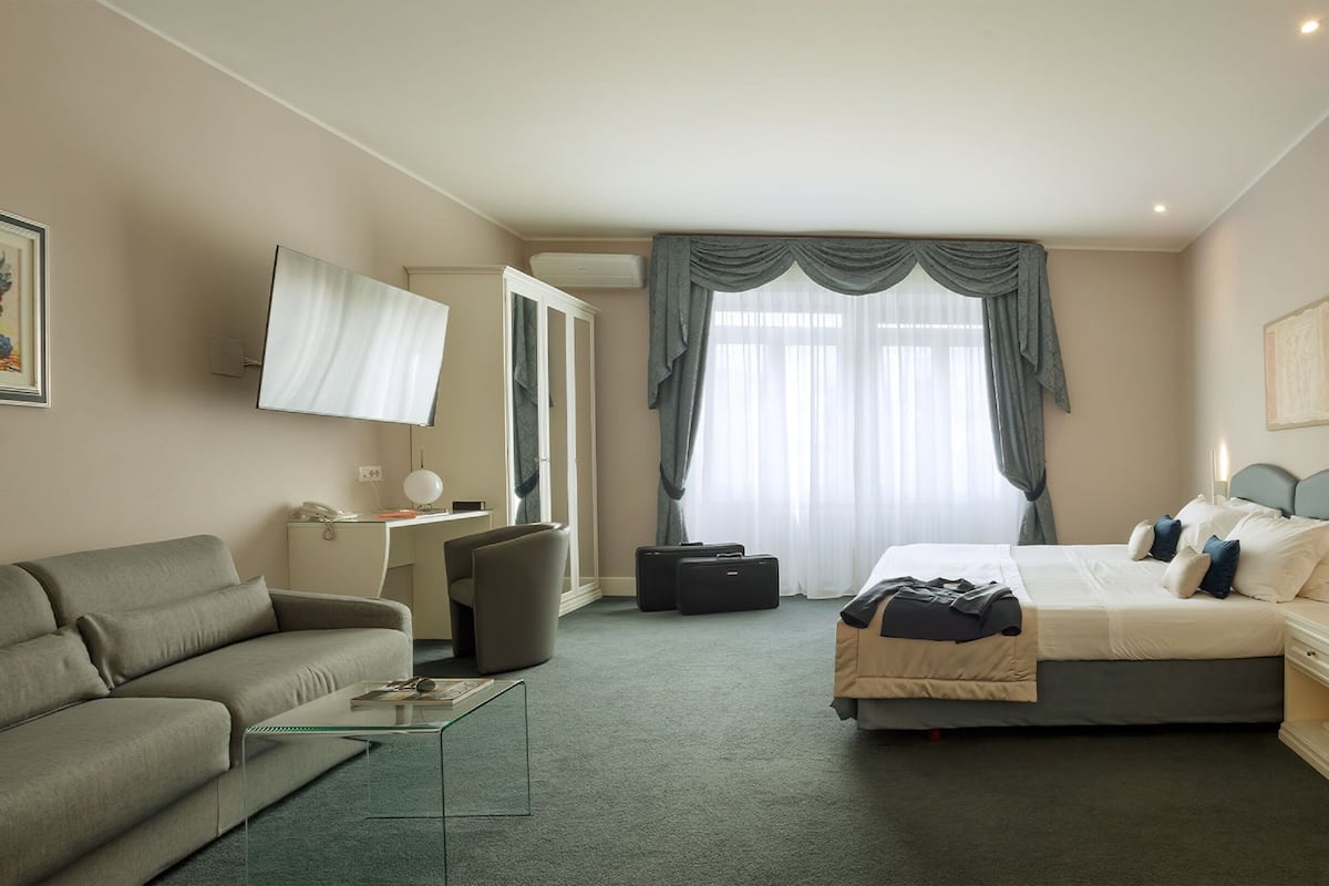 A hotel room with a bed, a sofa, a desk with a lamp, a chair, and a large window with curtains.