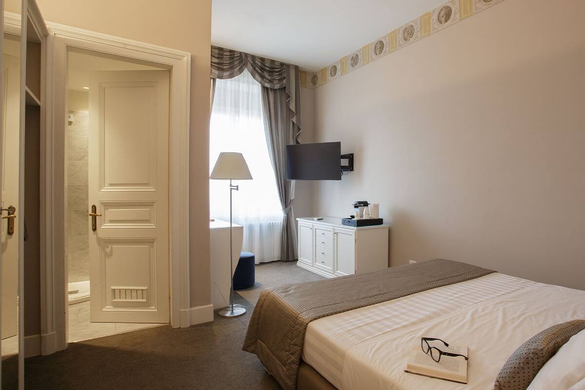 A hotel room with a bed, a desk with a flat-screen TV, a lamp, and a wardrobe.