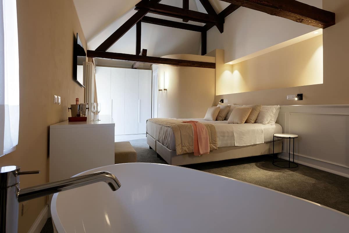 A modern bedroom with a large white bathtub, a bed with beige bedding, and a small bedside table.
