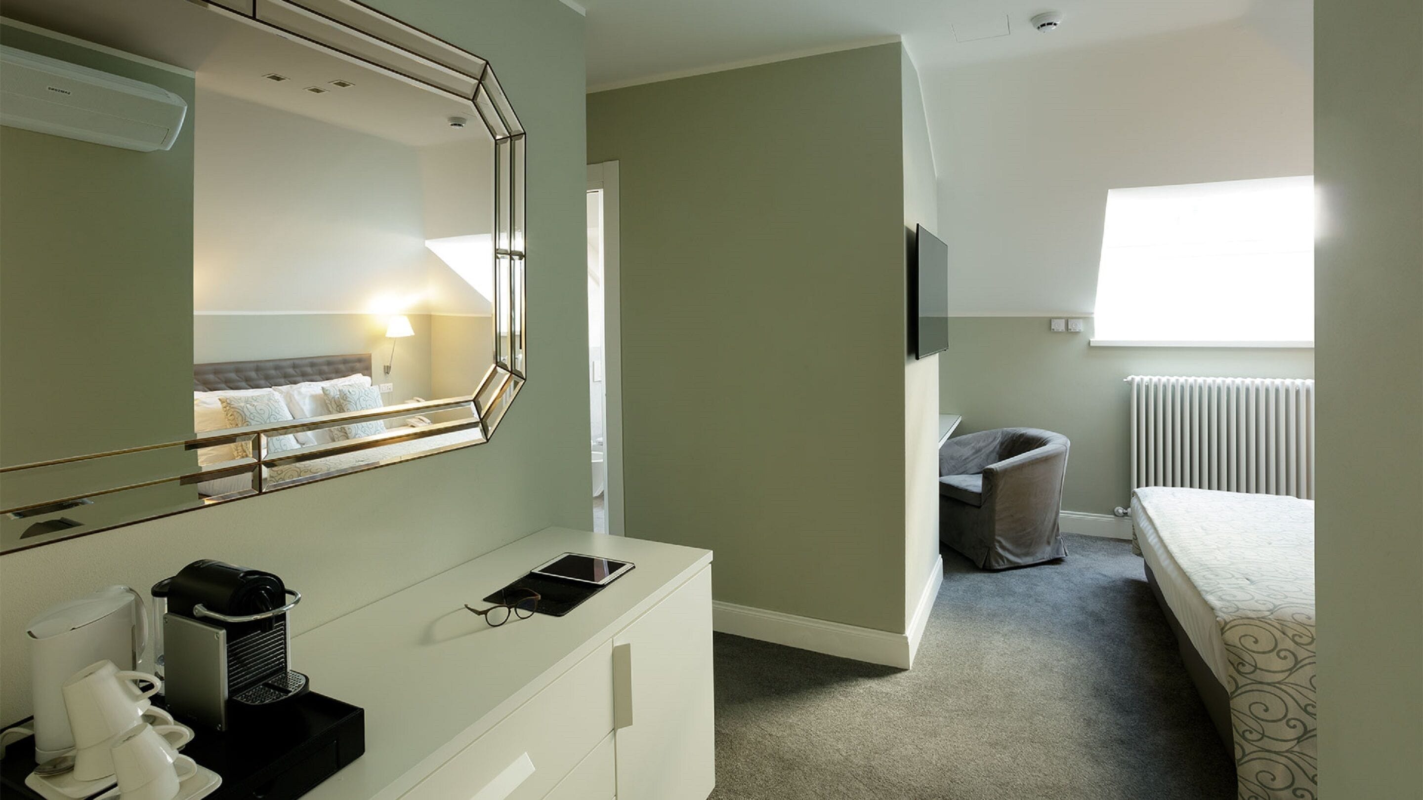 Une chambre d’hôtel avec un lit, un bureau avec un miroir, un radiateur et une fenêtre.