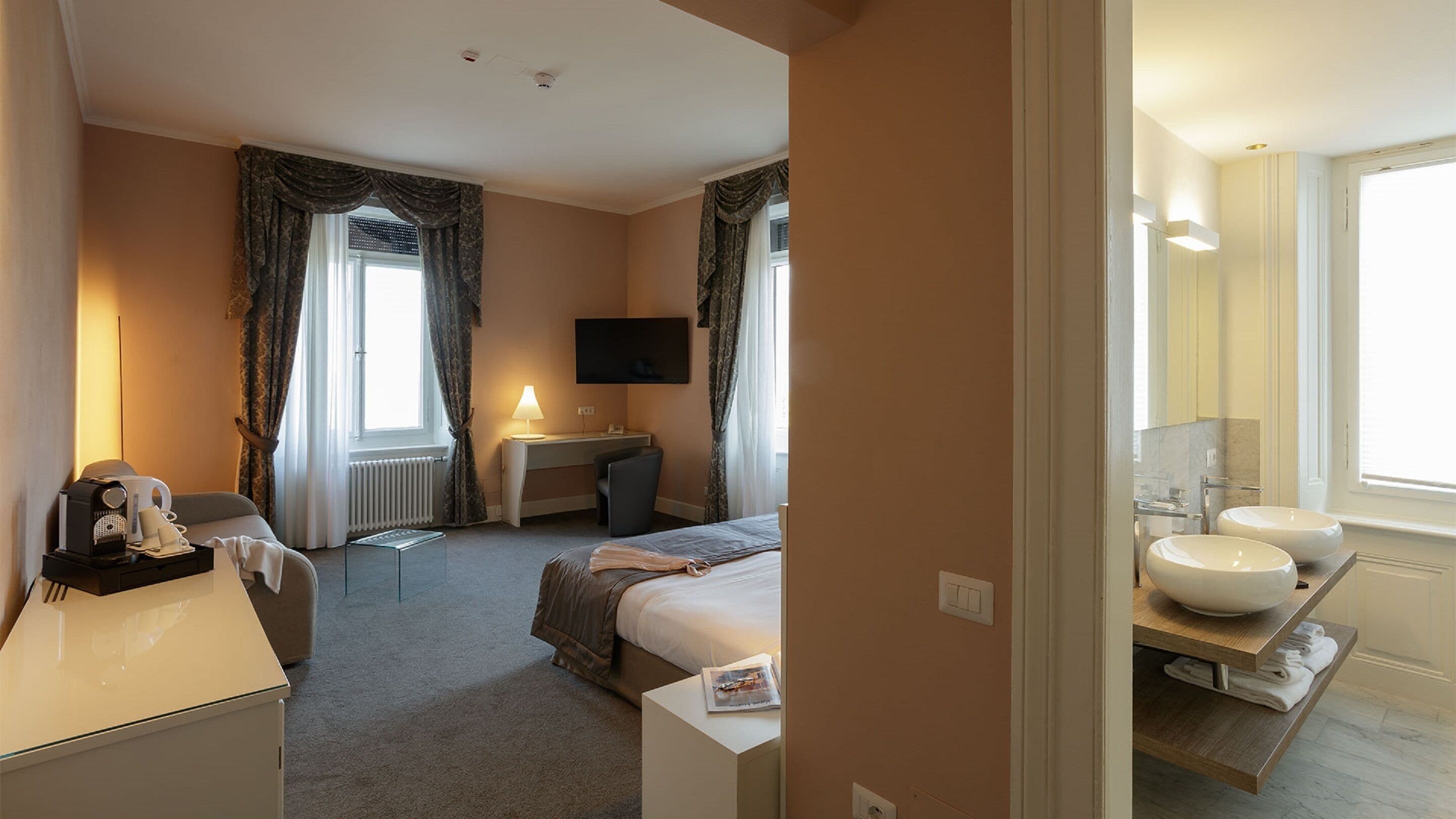 Habitación de hotel con cama, escritorio, televisor, lámpara y baño con lavamanos y espejo.