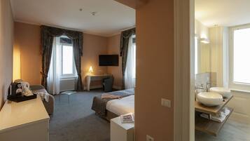 Habitación de hotel con cama, escritorio, televisor, lámpara y baño con lavamanos y espejo.