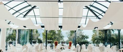 An indoor venue with round tables set for a formal event, featuring white tablecloths and chairs, and a glass ceiling structure.