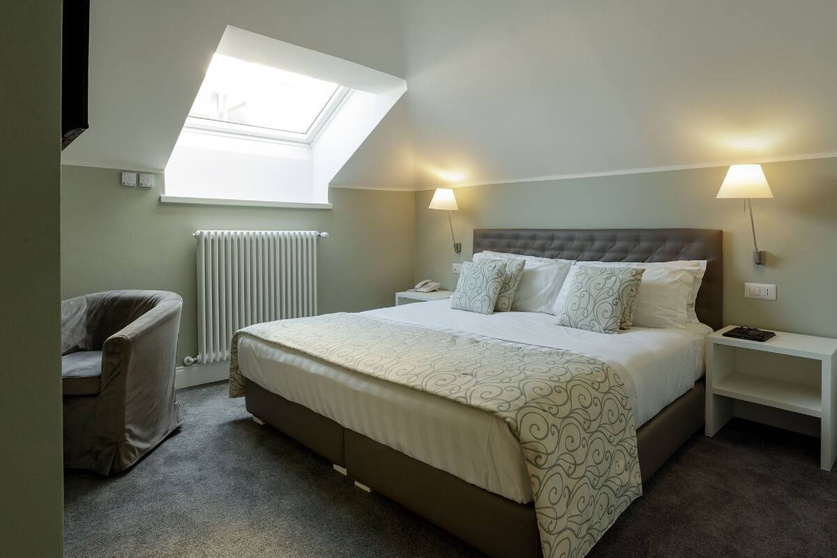 A bedroom with a bed, a chair, a nightstand, a lamp, a radiator, and a skylight.