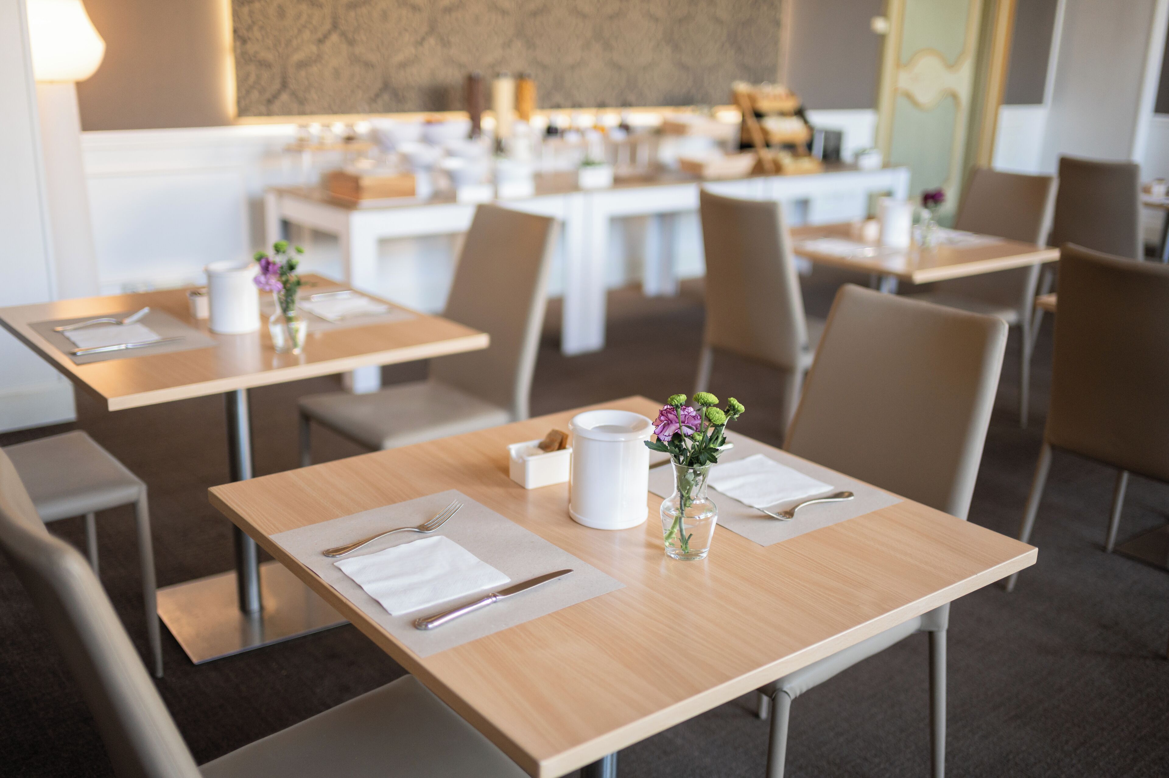 A modern dining area with neatly arranged tables, each set with cutlery, napkins, and a small vase with flowers.