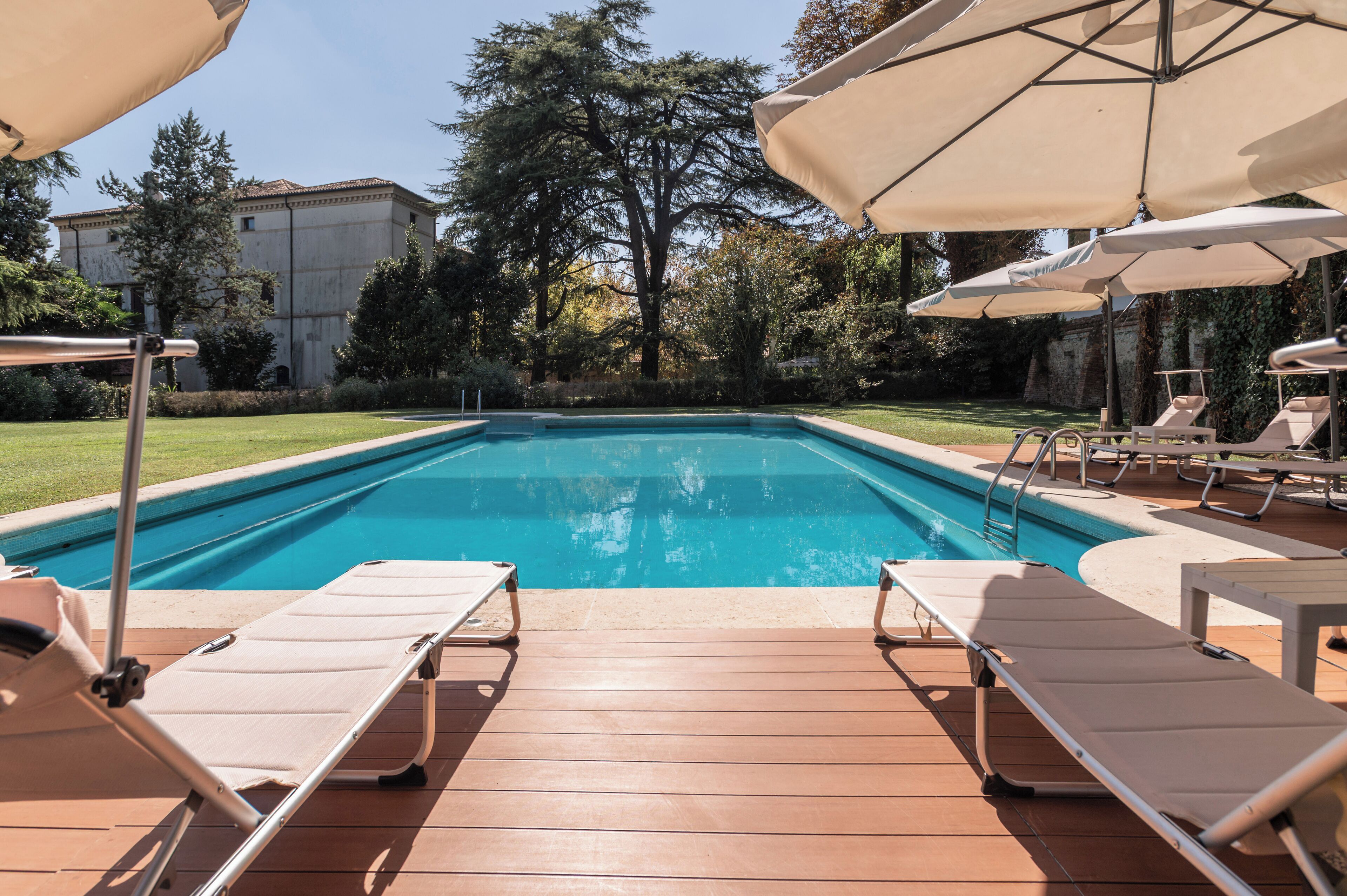Una piscina con sillas de descanso y sombrillas, rodeada de vegetación y un edificio al fondo.
