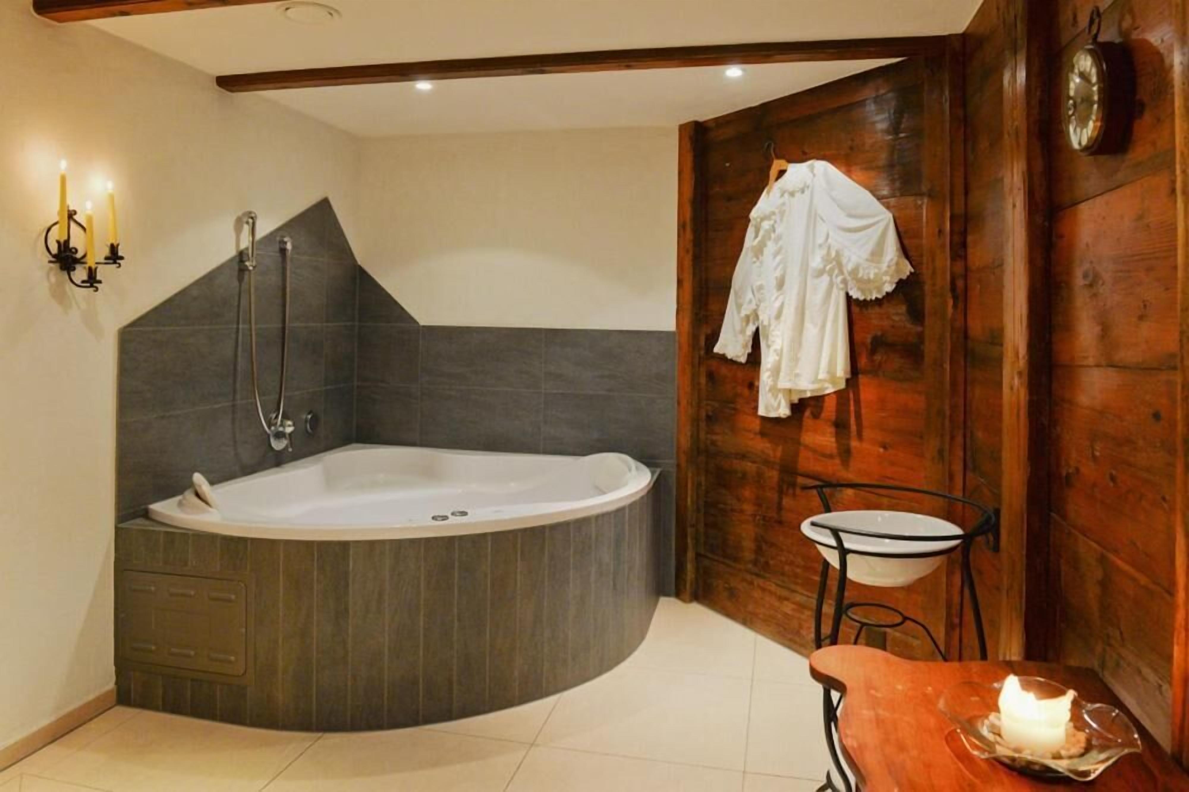 A modern bathroom with a large bathtub, a wooden wall, a small table with a candle, and a coat hanging on the wall.