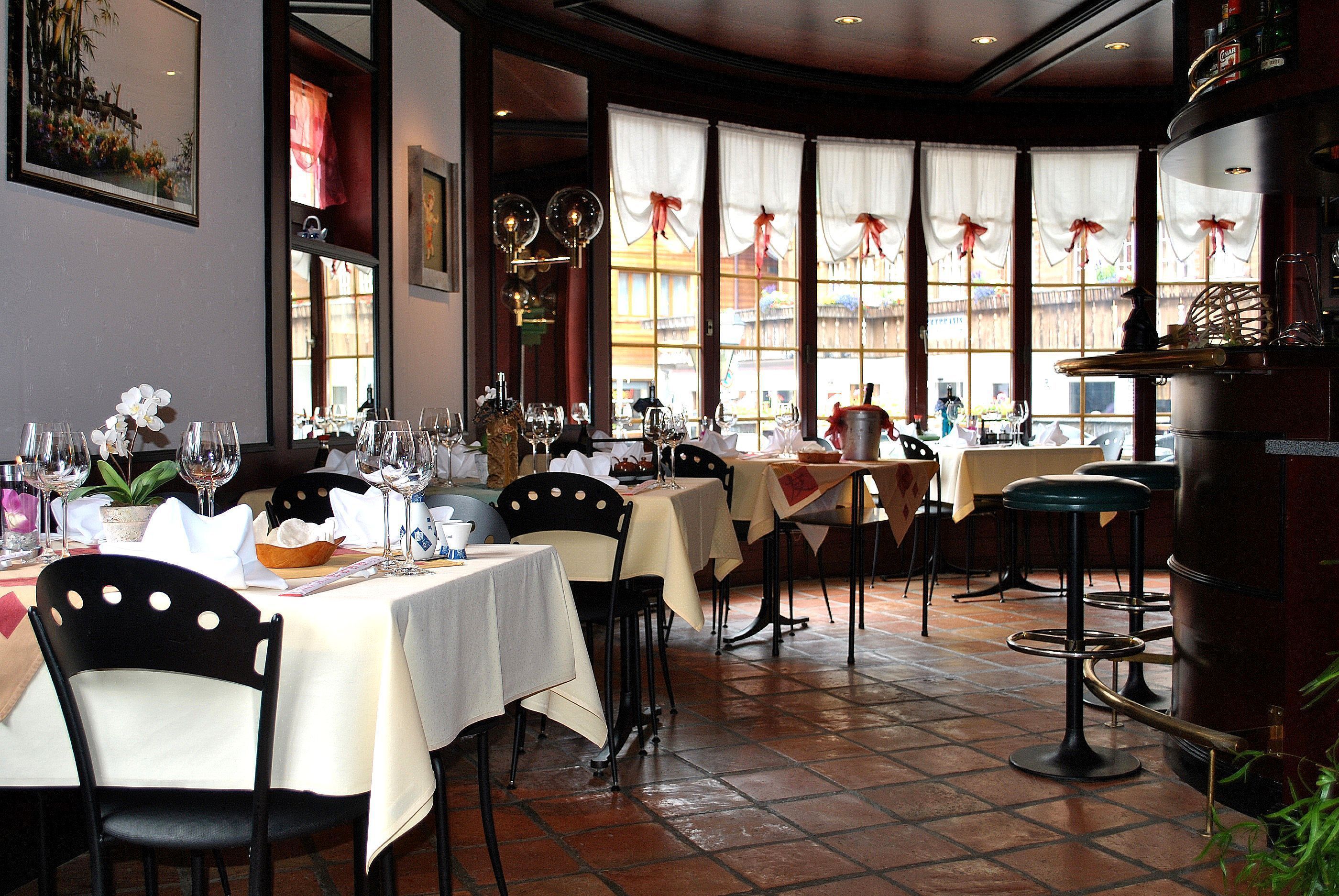 A restaurant with multiple tables and chairs, large windows with curtains, and a tiled floor.