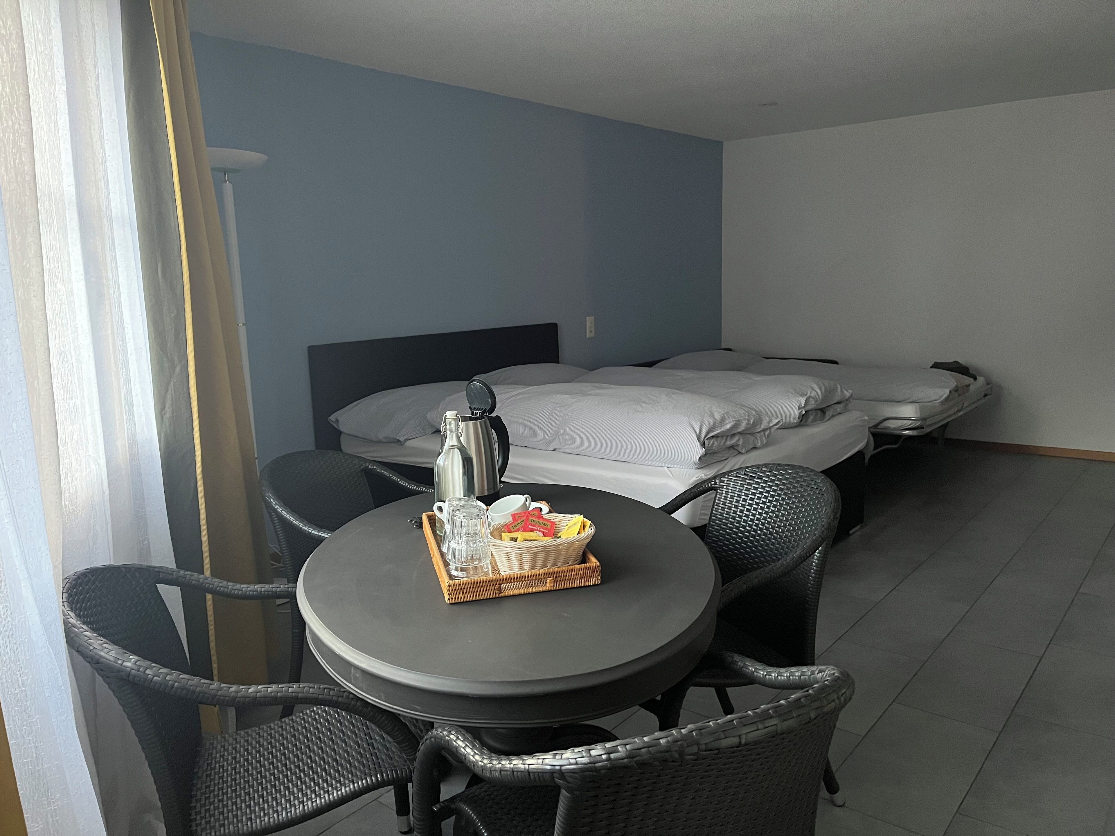 Een hotelkamer met een bed, een ronde tafel met stoelen, een koffiezetapparaat en een mand met brood op de tafel.