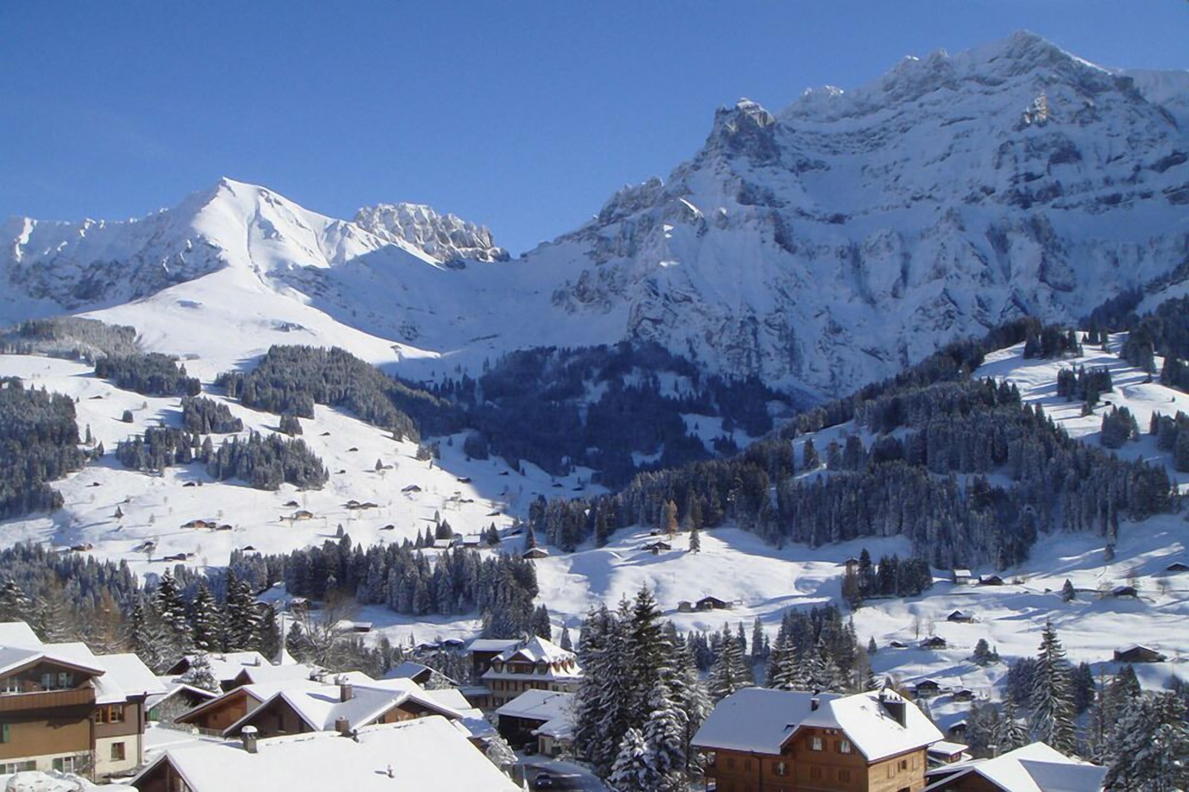 A snow-covered mountain landscape with a ski resort in the foreground, featuring chalet-style buildings and a clear view of the peaks.