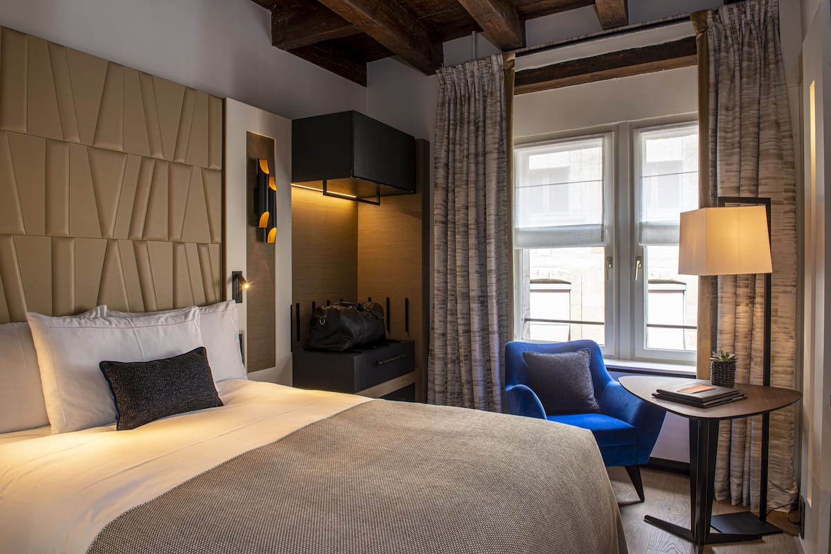 A hotel room with a large bed, a blue armchair, a small table with a lamp, and a view of the outside through a window with curtains.
