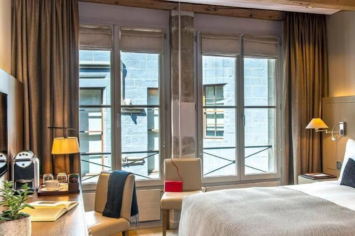 A hotel room with a large bed, a chair, a small table, a lamp, and a view of the outside.