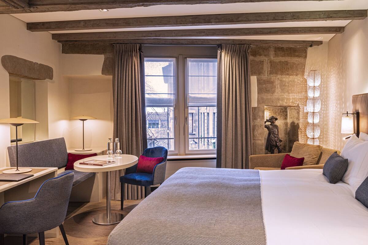 A hotel room with a large bed, a desk with a chair, a small round table, and a balcony with curtains.