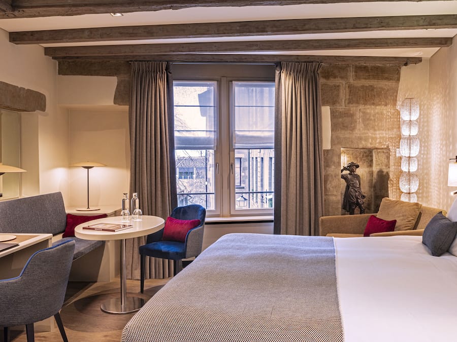 Habitación de hotel con una cama grande, un escritorio con silla, una mesita redonda y un balcón con cortinas.