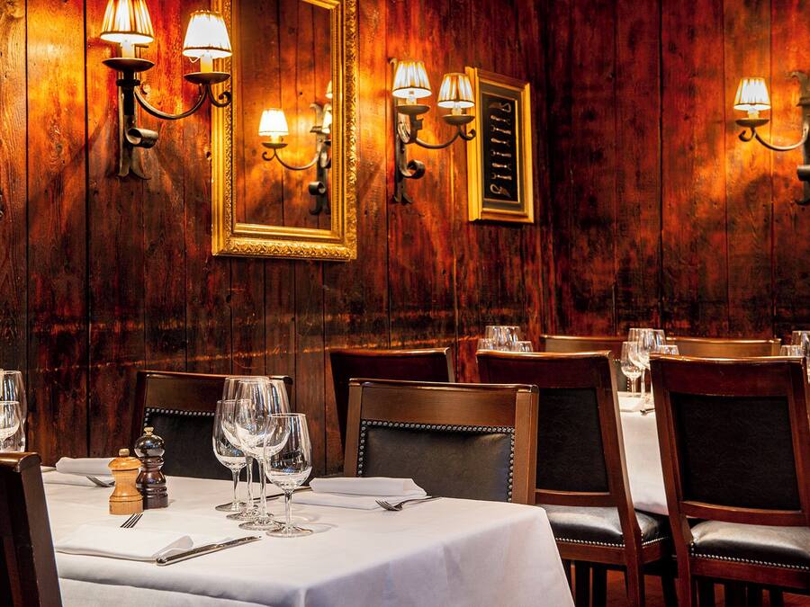 En restauranginteriör med väggar beklädda med träpaneler, kristallkronor och bord som är dukade med vinglas och bestick.