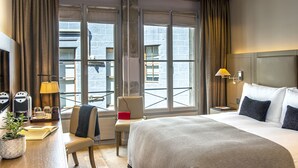 A hotel room with a large bed, a desk with a lamp, a chair, and a view of the city through large windows. - Hôtel Les Armures (Geneva)