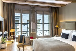 Quarto de hotel com uma cama grande, uma escrivaninha com luminária, uma cadeira e vista para a cidade através de amplas janelas.