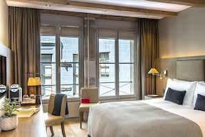 A hotel room with a large bed, a desk with a lamp, a chair, and a view of the city through large windows. - Hôtel Les Armures (Geneva)