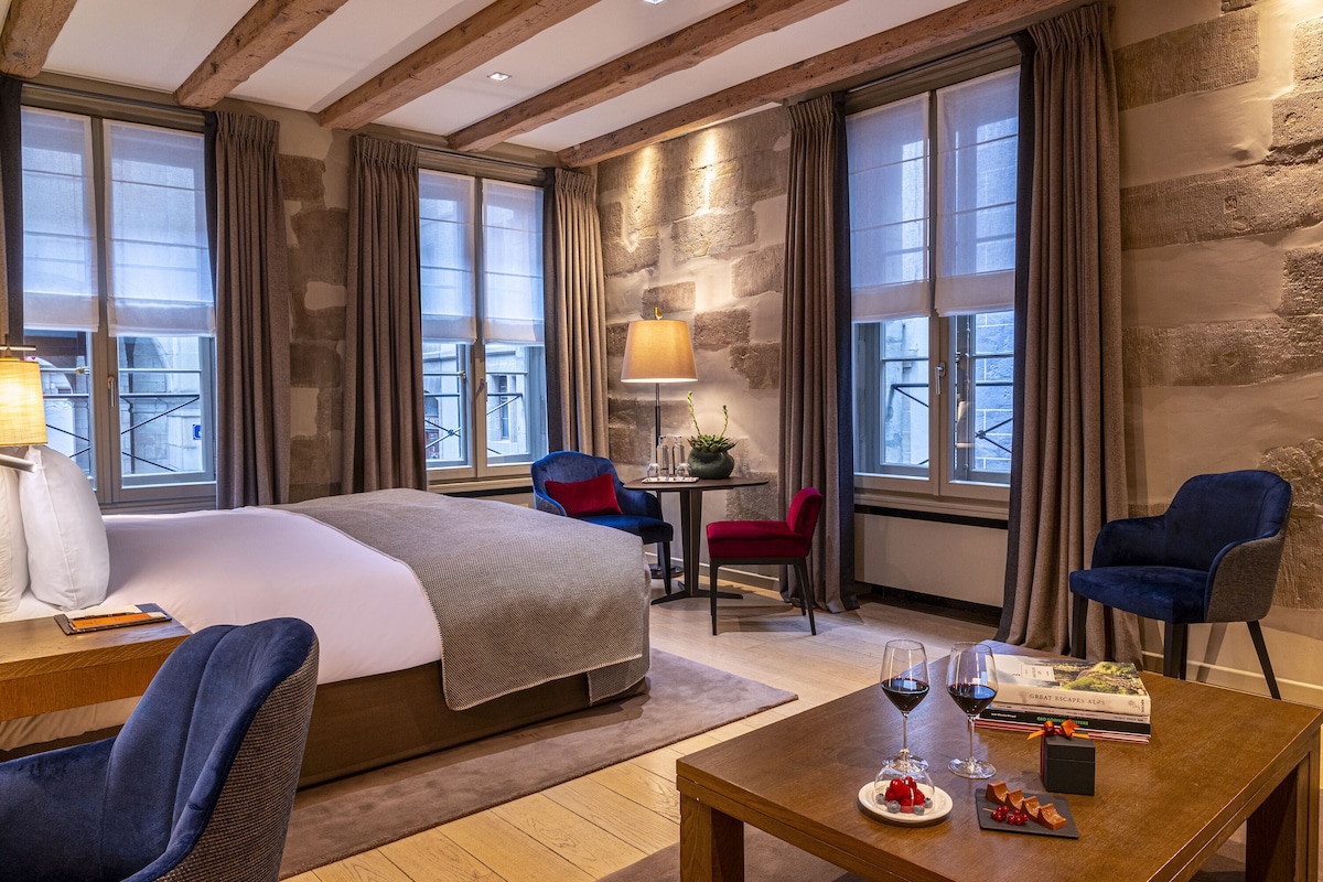 Une chambre d’hôtel avec un grand lit, deux fauteuils, une petite table avec un verre à vin et un livre, et une vue sur la ville à travers de grandes fenêtres.