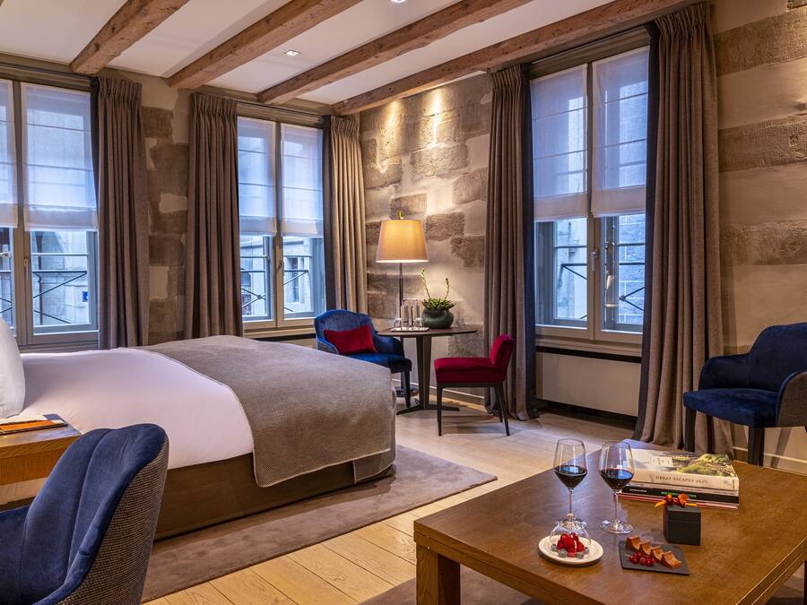Habitación de hotel con una cama grande, dos sillones, una mesita con una copa de vino y un libro, y vistas a la ciudad a través de amplios ventanales.