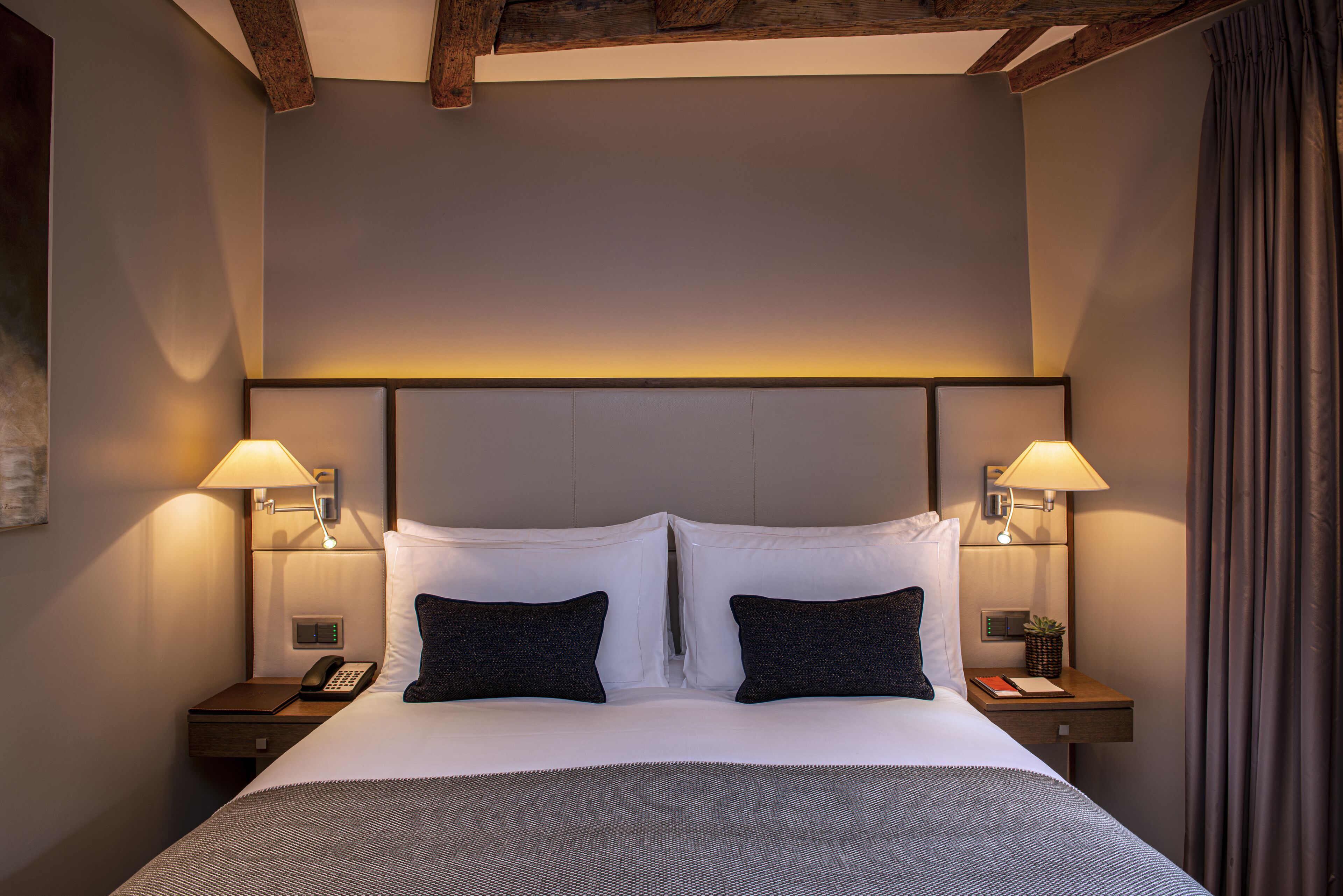 A neatly made bed with white and dark pillows, two bedside lamps, a nightstand with a phone, and a small plant.