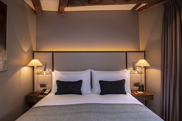 A neatly made bed with white and dark pillows, two bedside lamps, a nightstand with a phone, and a small plant.