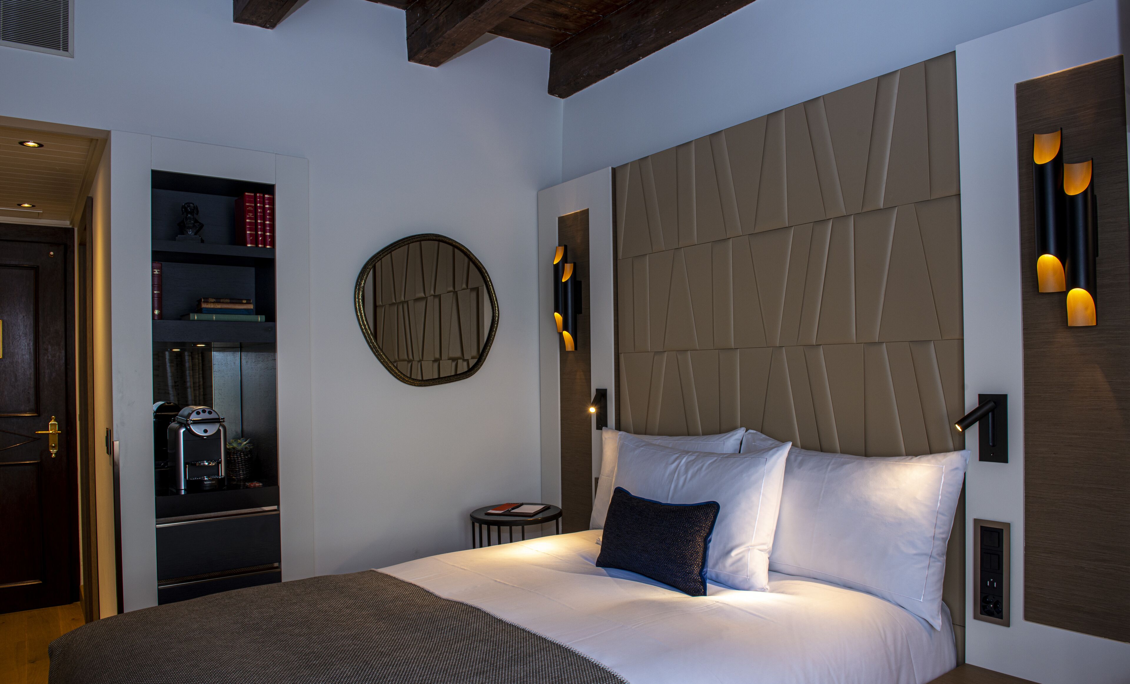 A modern hotel room with a large bed, a round mirror, a small side table, and a built-in bookshelf.