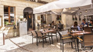 An outdoor cafe area with tables and chairs, a stone building with a sign reading "RESTAURANT LES ARMURES", and potted plants.
