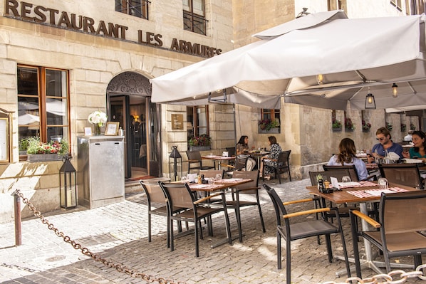Un espace café en plein air avec des tables et des chaises, un bâtiment en pierre portant une enseigne indiquant « RESTAURANT LES ARMURES », et des plantes en pot.