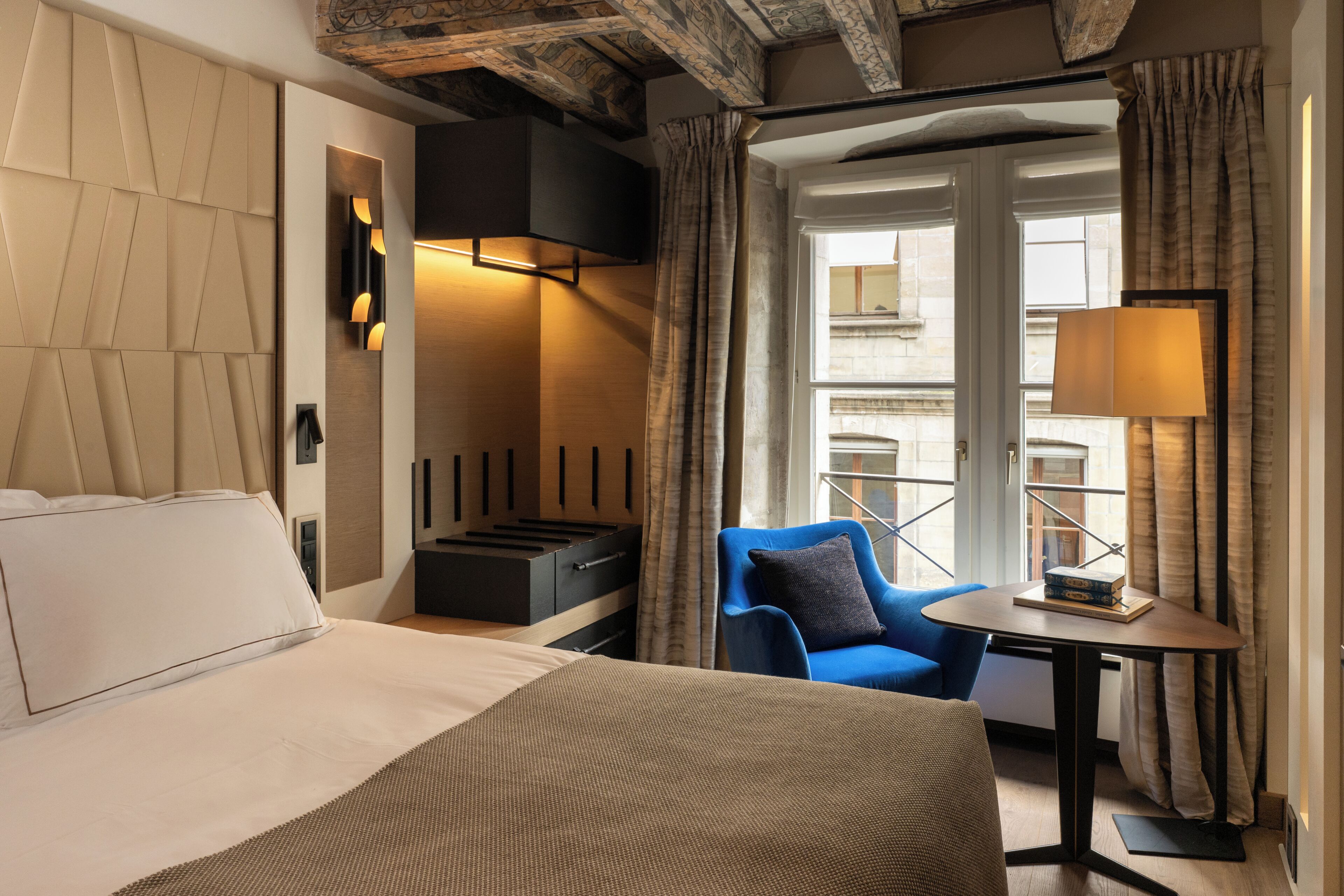 A hotel room with a bed, a seating area with a blue chair, a small table, and a window with curtains.