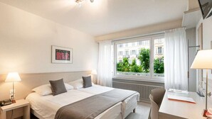 A hotel room with a bed, bedside table with lamp, desk with chair, and a window with curtains. - Hotel des Arts (Neuchatel)