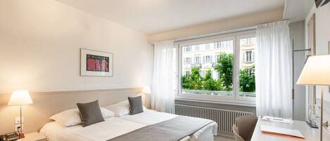 A hotel room with a bed, bedside table with lamp, desk with chair, and a window with curtains.