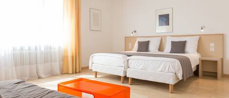 A modern hotel room with two beds, a bright window with sheer curtains, and a small orange table.