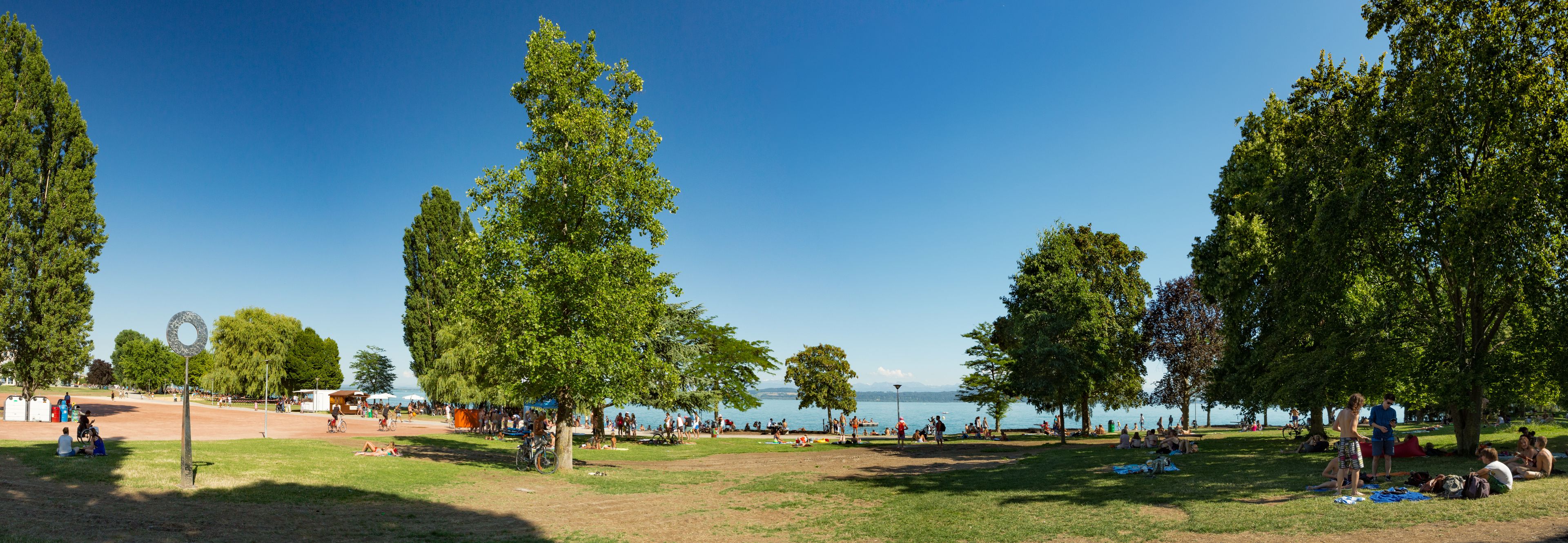 Un parque con zonas verdes, árboles y un lago al fondo.