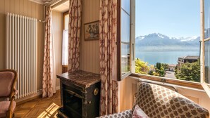 A room with a view of a lake and mountains, a wooden chair, a radiator, and a vintage stove. - Hôtel Masson (Veytaux)