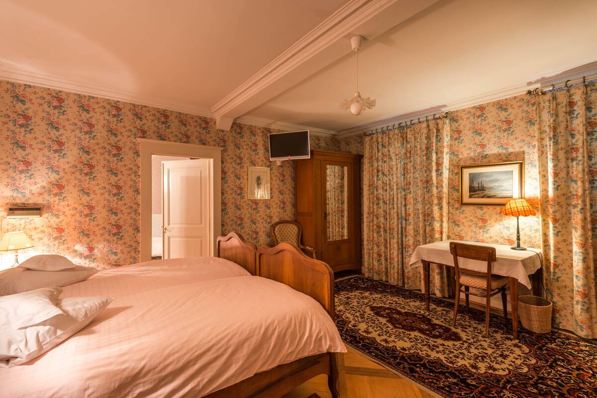 A bedroom with a large bed, a desk with a lamp, a chair, a television, and a door.