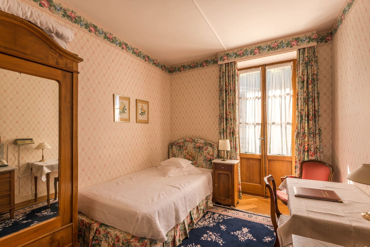 A bedroom with a single bed, a wooden wardrobe, a desk with a chair, a small table, and a window with floral curtains.