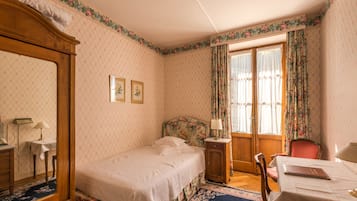 A bedroom with a single bed, a wooden wardrobe, a desk with a chair, a small table, and a window with floral curtains.