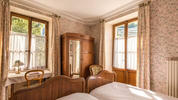 A bedroom with two wooden beds, a wooden wardrobe, a small table with a lamp, and floral-patterned curtains.