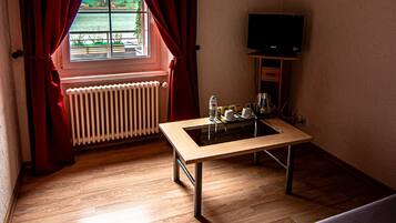 Una habitación con suelo de madera, una pequeña mesa con cafetera y tazas, un radiador, un televisor sobre un mueble y una ventana con cortinas.