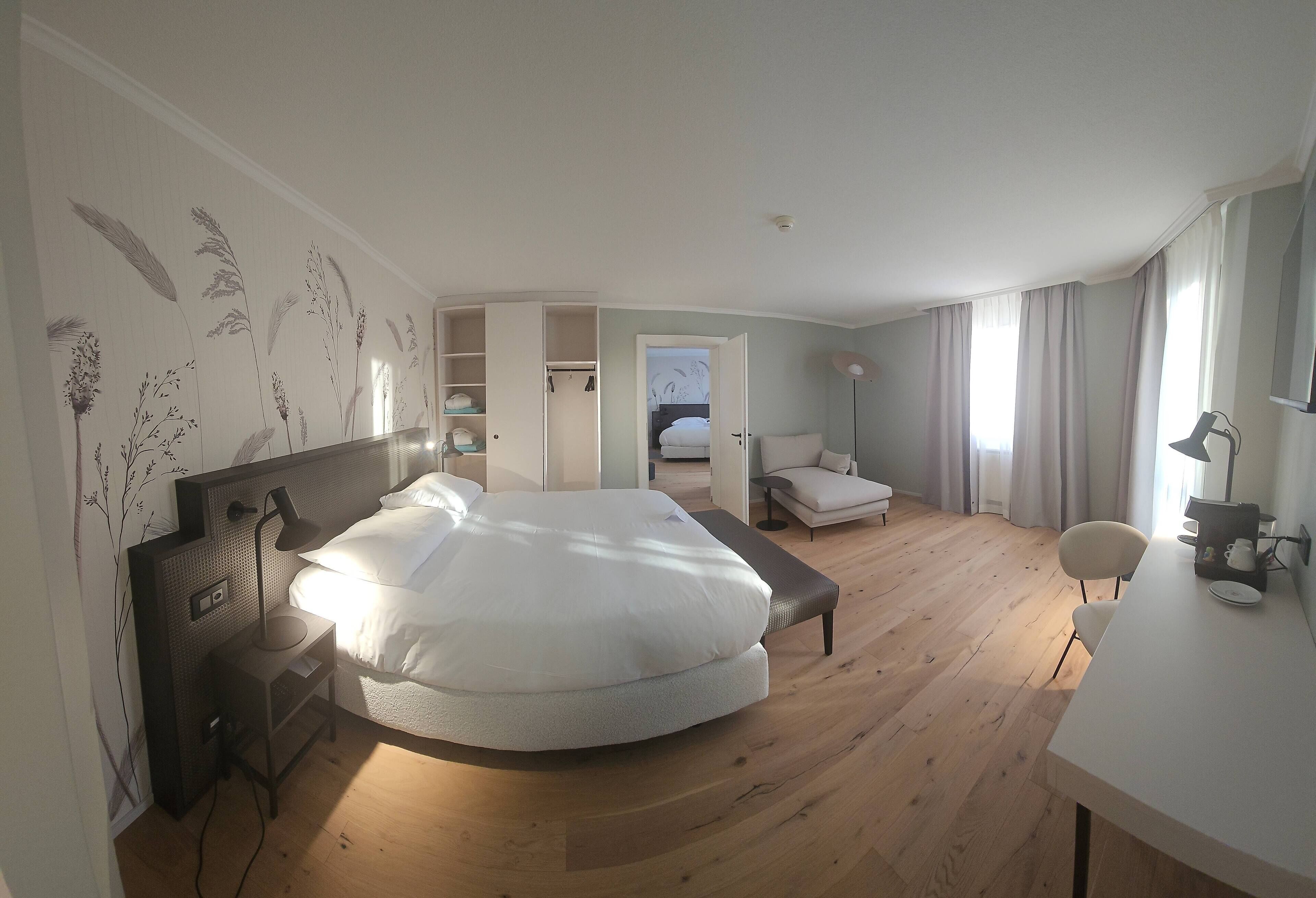 A modern bedroom with a large bed, a desk, and a chair. There is a wardrobe and a door leading to another room.