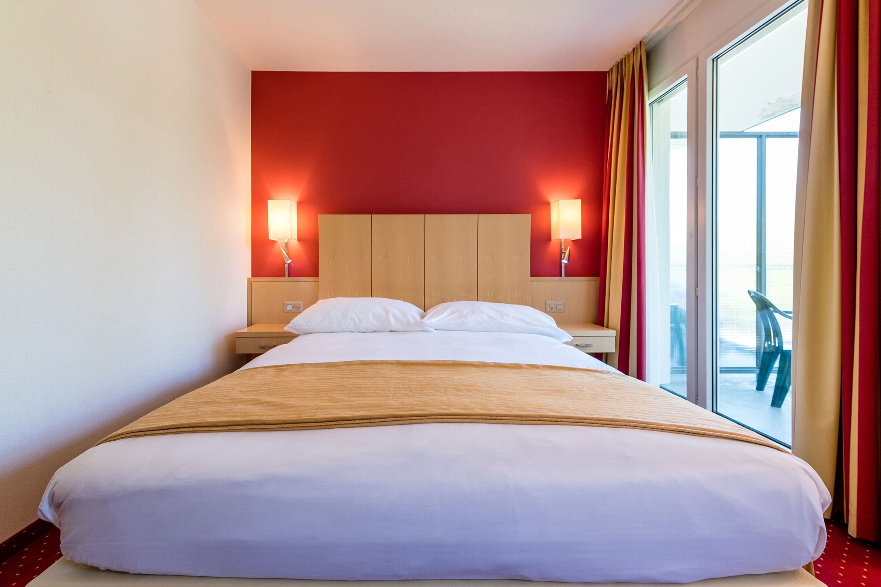 Une chambre d’hôtel avec un grand lit, deux lampes de chevet, une tête de lit en bois et une vue sur l’extérieur grâce à une grande fenêtre.
