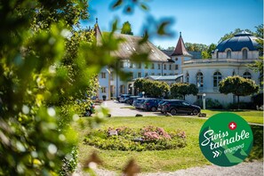 A historic building with a large dome and multiple windows, surrounded by a well-maintained garden and parked cars. - Grand Hôtel et Centre Thermal (Yverdon-les-Bains)
