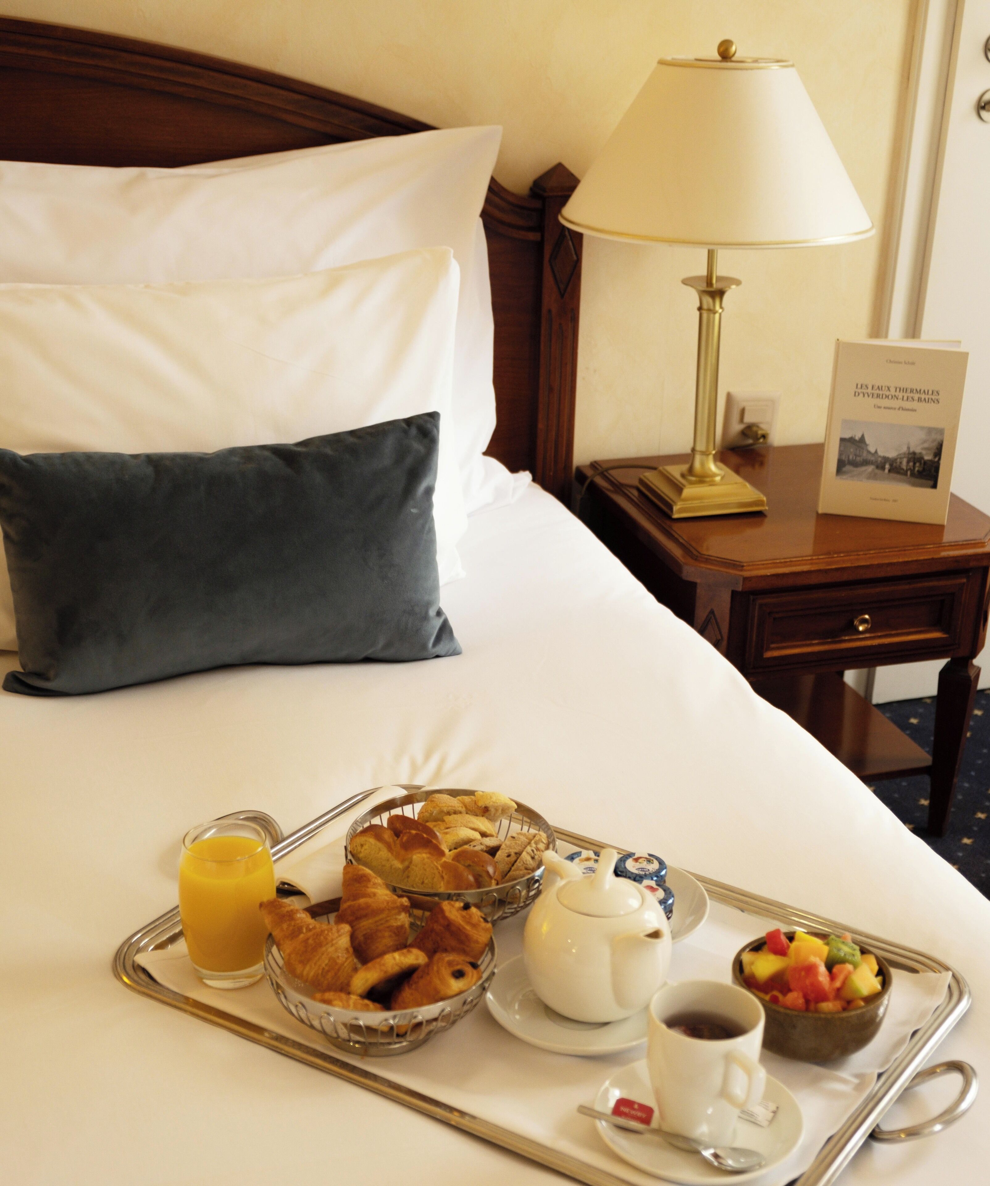A neatly made bed with white and dark pillows, a bedside table with a lamp and a book, and a tray with breakfast items including croissants, fruit, coffee, and juice.