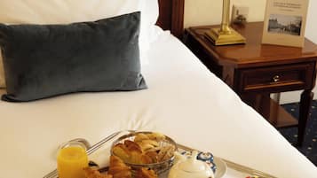 A neatly made bed with white and dark pillows, a bedside table with a lamp and a book, and a tray with breakfast items including croissants, fruit, coffee, and juice.