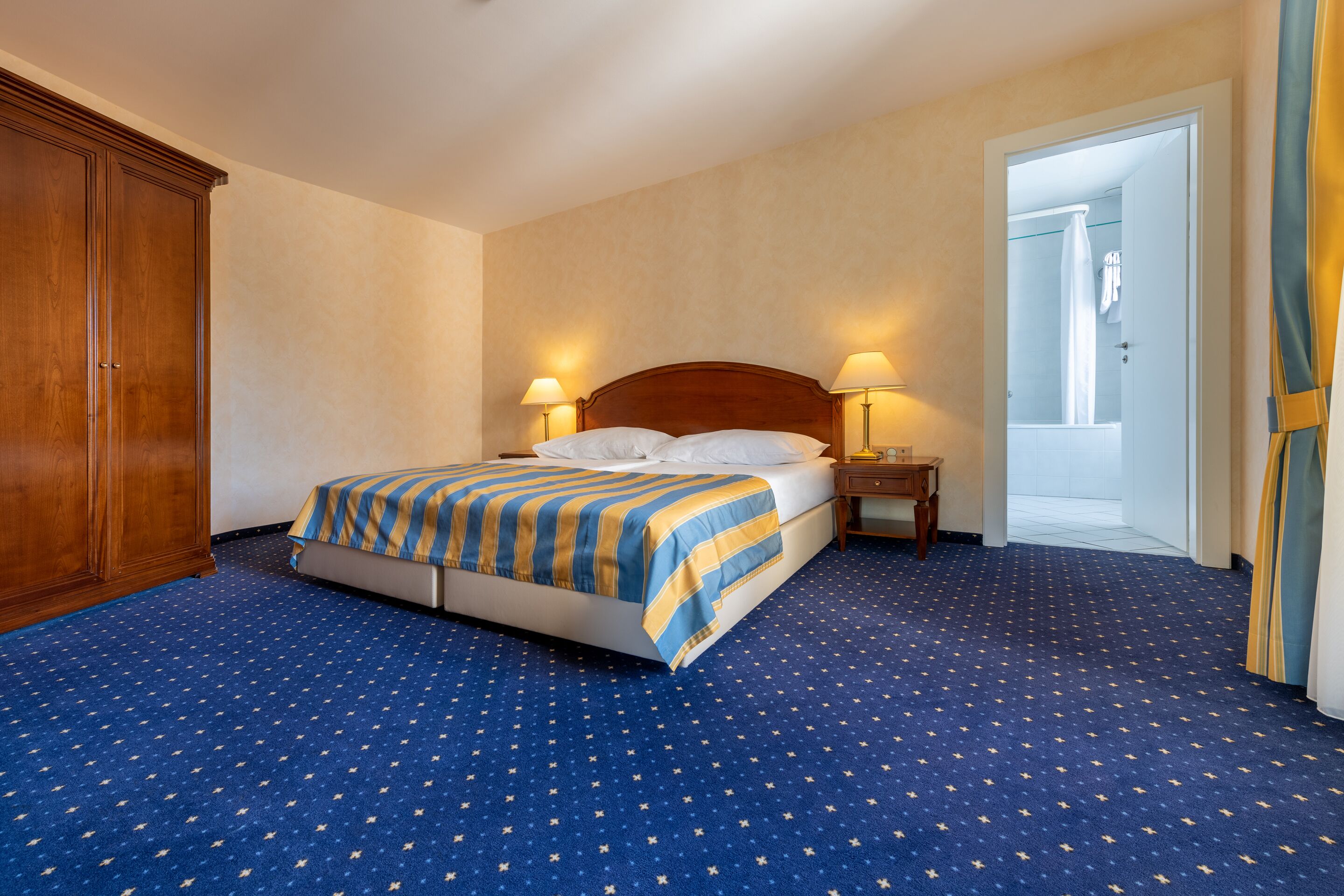 Habitación de hotel con una cama grande, dos mesitas de noche, un armario y un baño visible a través de una puerta abierta.