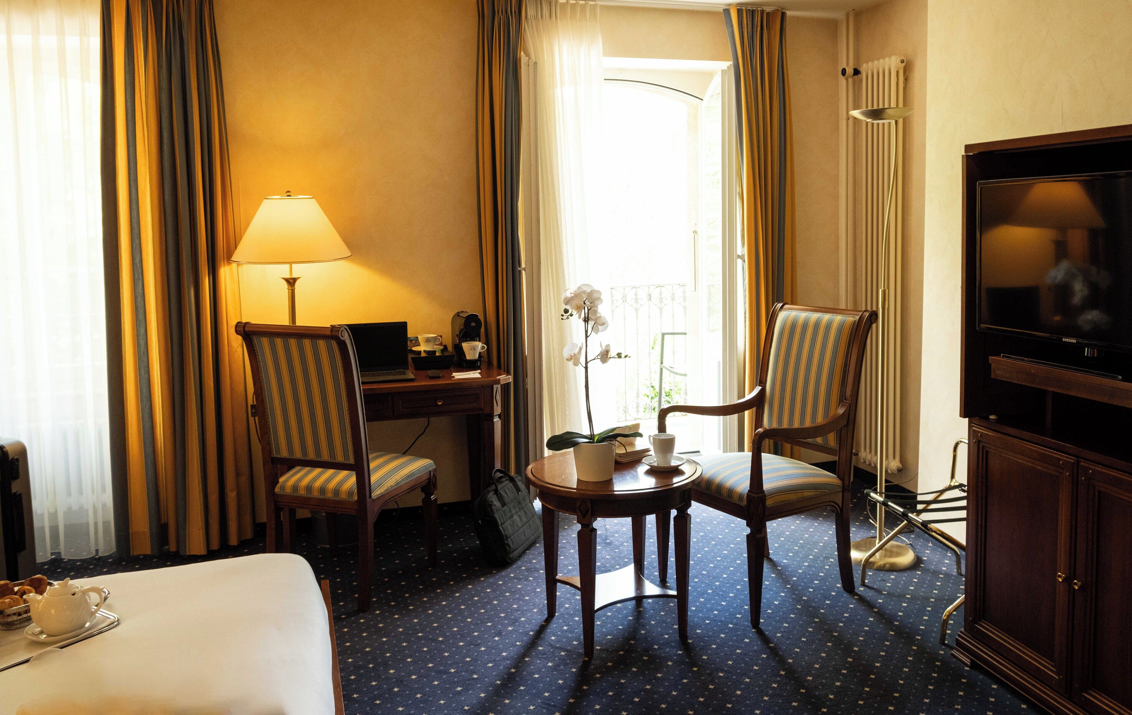 Et hotelværelse med en seng, et skrivebord med lampe, to stribede stole, et lille bord med en orkidé i potte, et fjernsyn og en radiator.