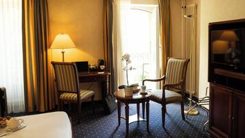 Une chambre d’hôtel avec un lit, un bureau avec une lampe, deux chaises à rayures, une petite table sur laquelle est posée une orchidée en pot, un téléviseur et un radiateur.